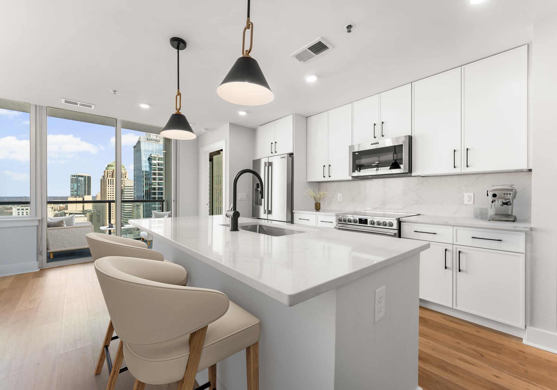 Modern white kitchen with island, pendant lights, bar stools, and city view through large windows.