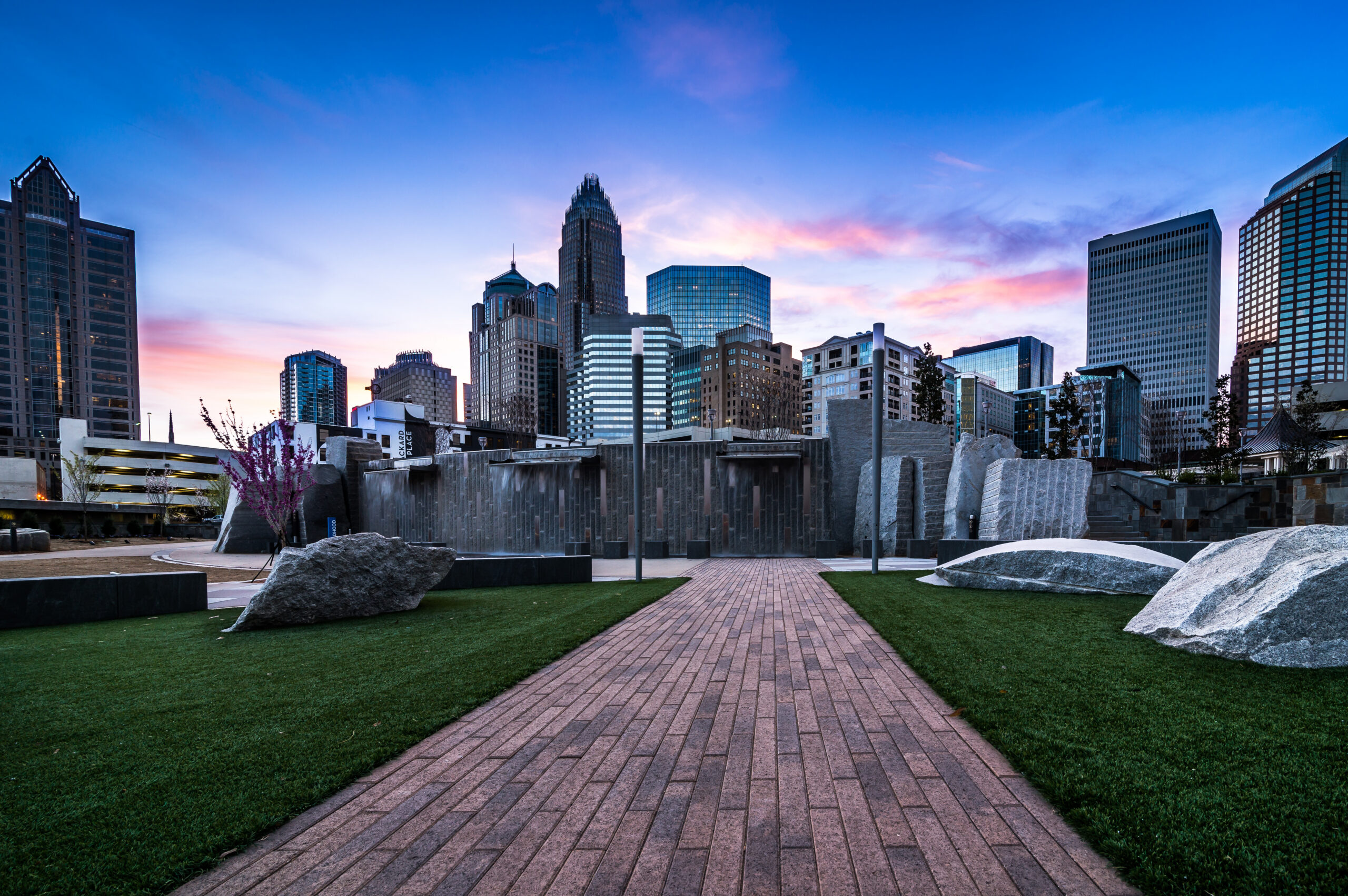 Brick pathway leads to urban park with modern buildings and skyscrapers at sunset, with green lawns and rocks in the foreground.