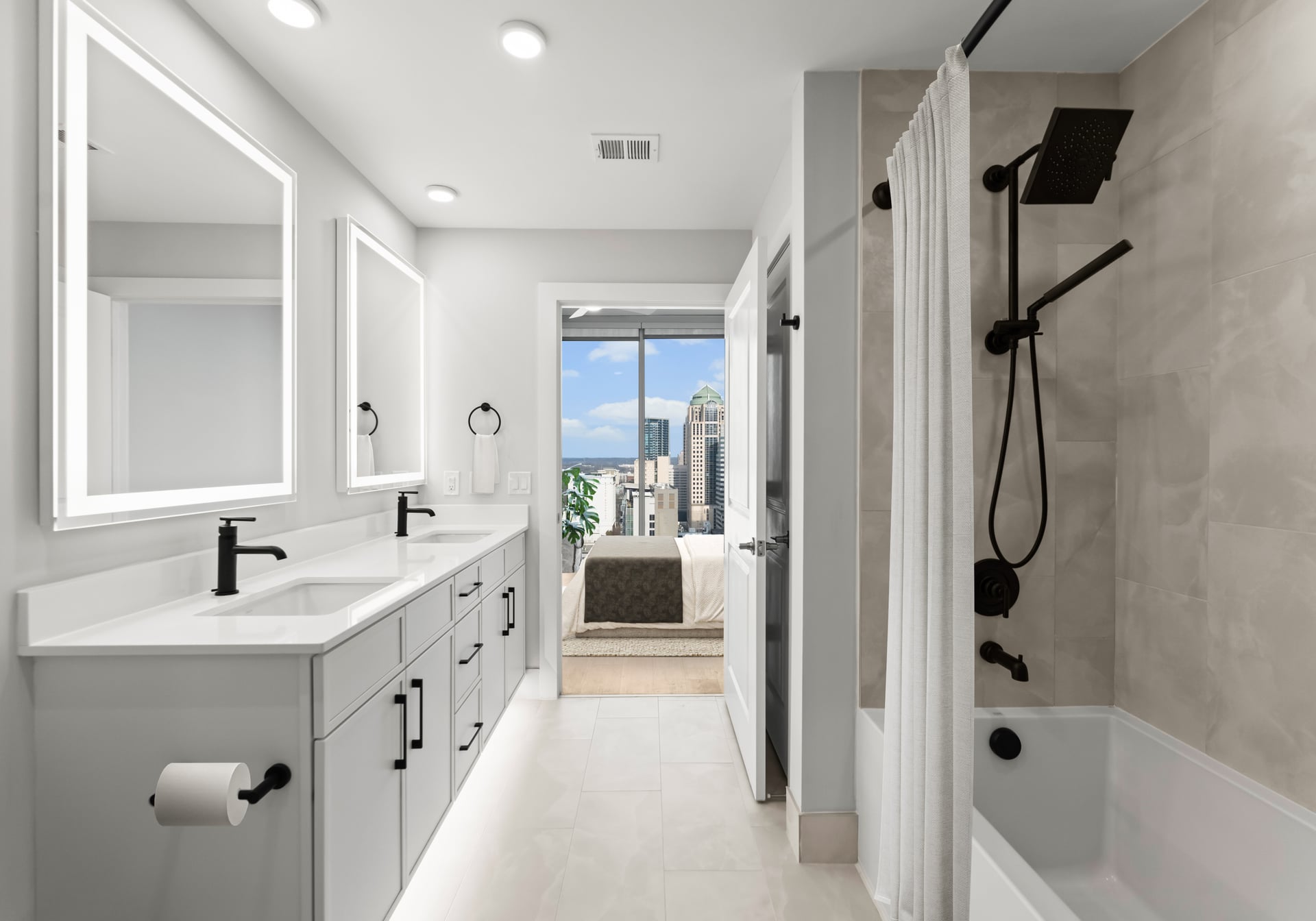 Modern bathroom with double sinks, lighted mirrors, and a shower-tub combo; bedroom and city view visible through the door.