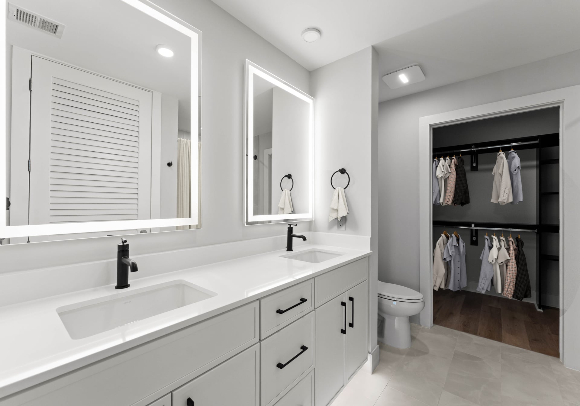 Modern bathroom with double sinks, white cabinets, illuminated mirrors, and open closet with hanging clothes in the background.