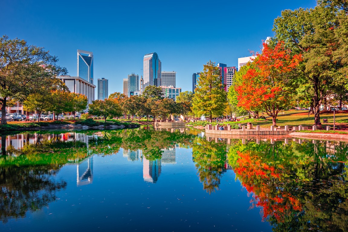 City skyline with modern buildings, colorful autumn trees, and clear reflections on a calm pond under a bright blue sky.