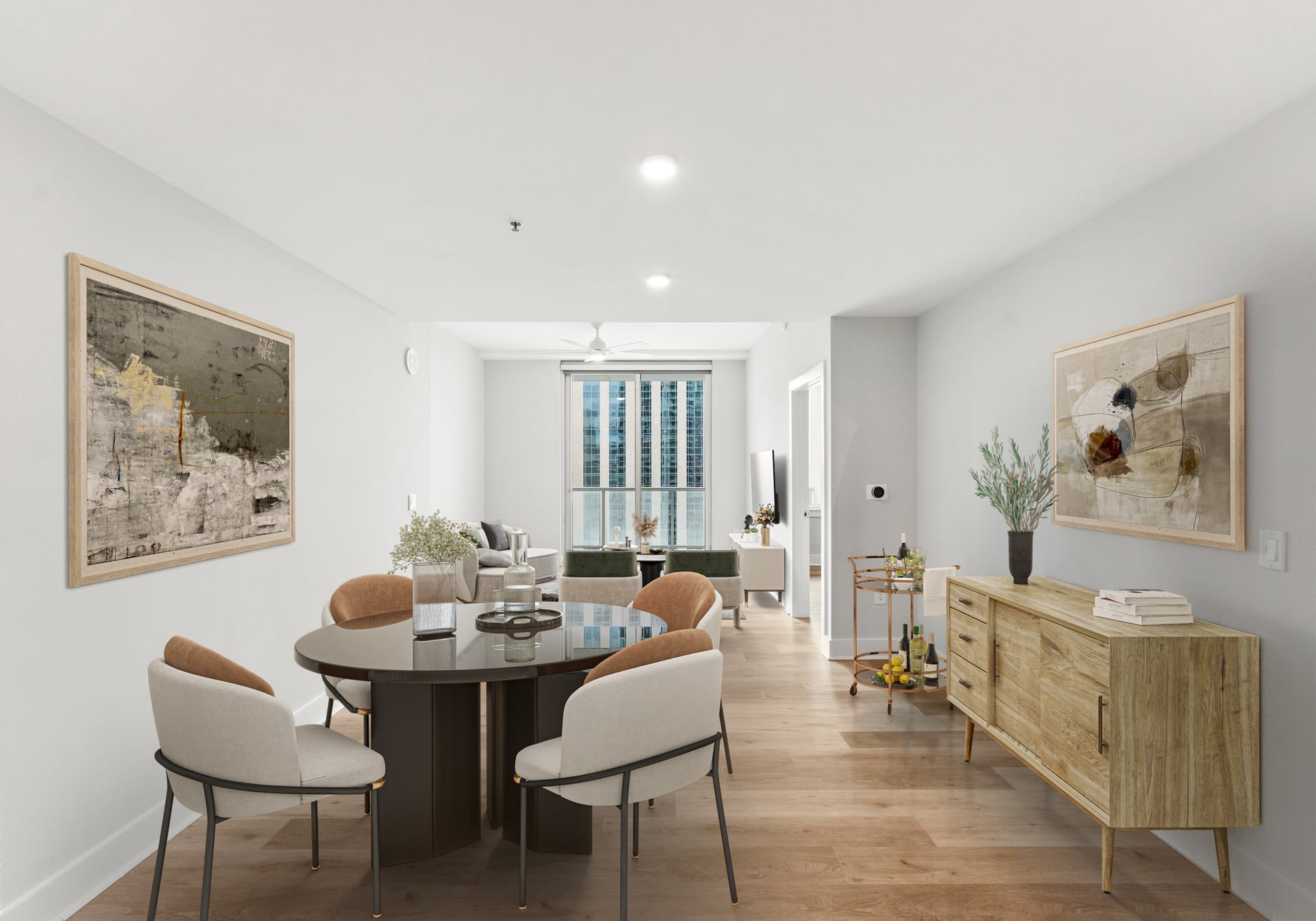 Modern living and dining area with round table, chairs, wooden sideboard, artwork, plants, and large window letting in natural light.