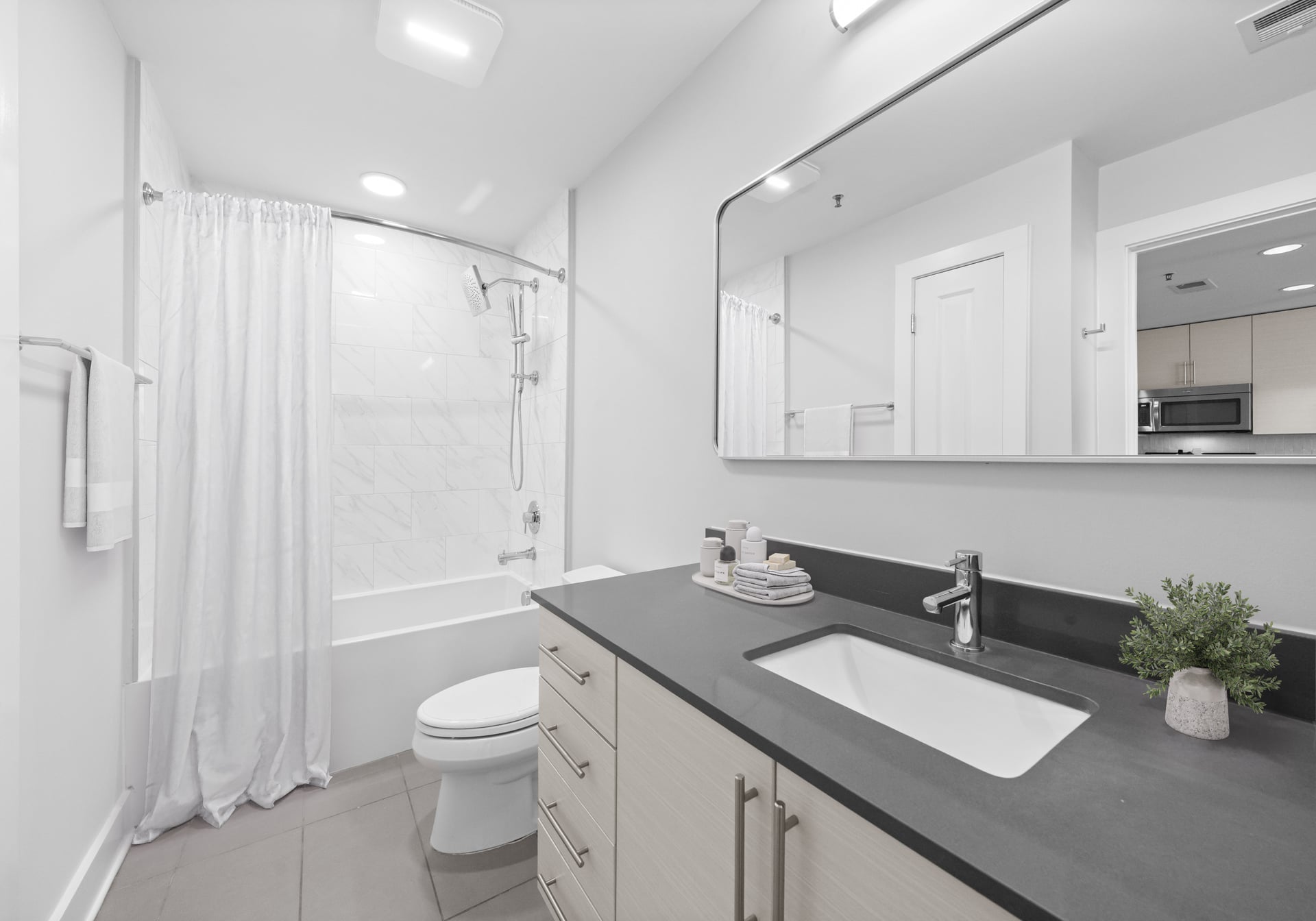 Modern bathroom with a bathtub, shower, toilet, sink, large mirror, and countertop with soap and a small plant.