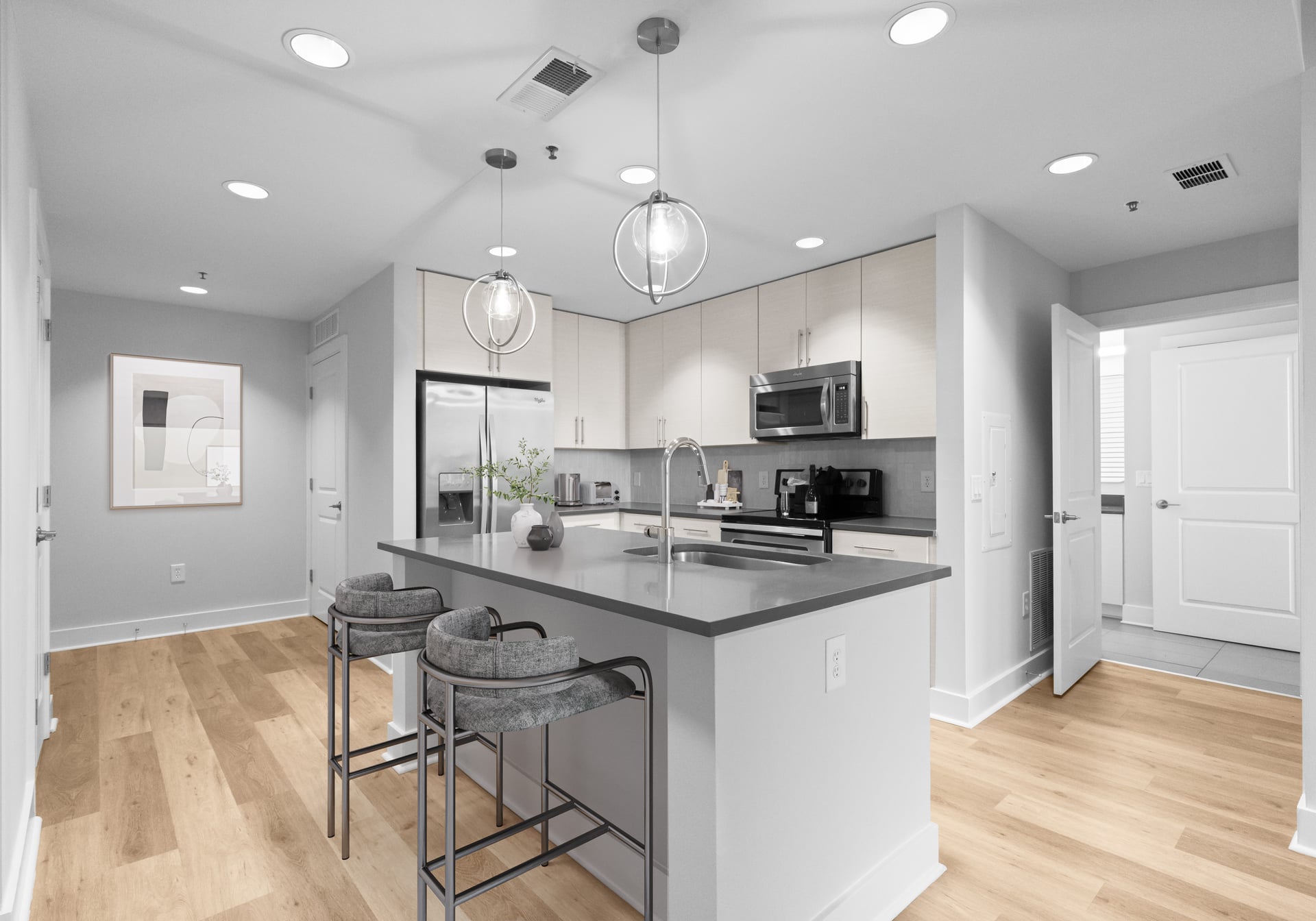 Modern kitchen with light wood floors, gray island, two barstools, stainless steel appliances, and pendant lights.