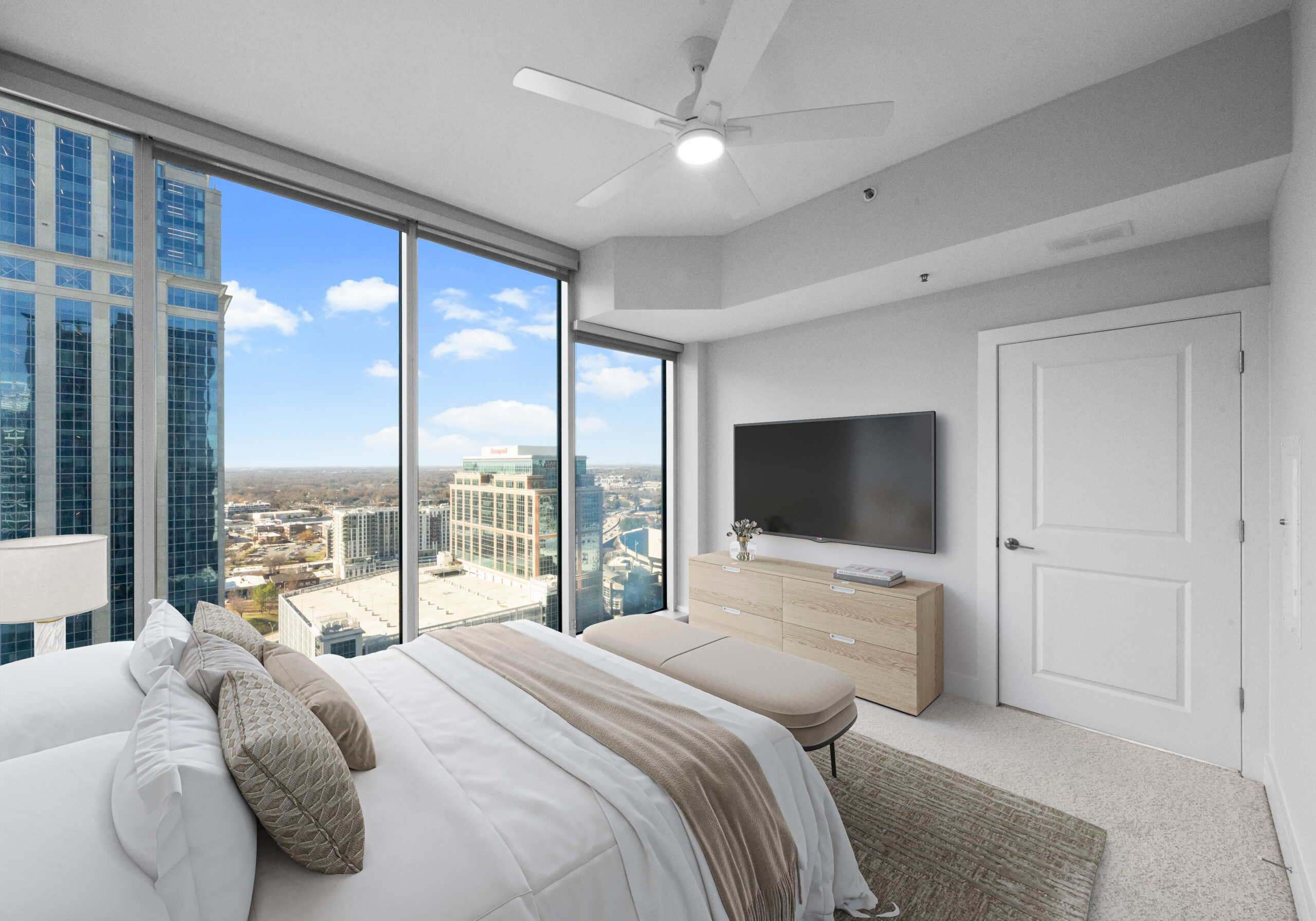 Modern living room with a gray sofa, wall art, TV, coffee table, lamp, and balcony view of city buildings through glass doors.