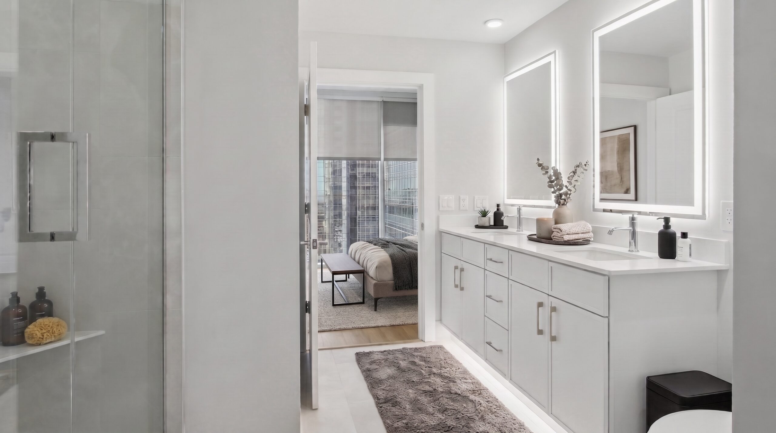 Modern bathroom with a double vanity and a glass shower, looking into a bedroom with city views.