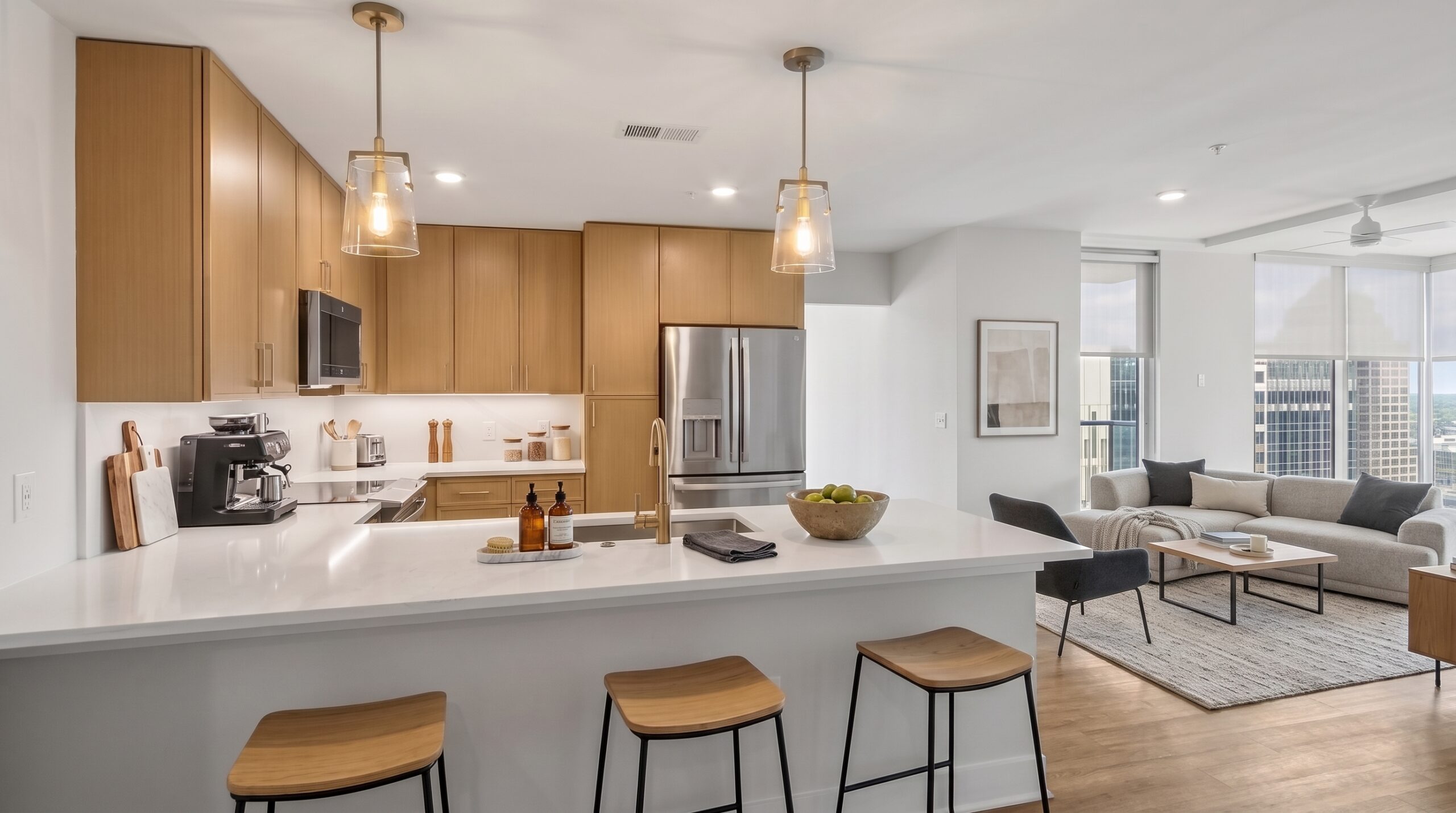 Modern kitchen with light wood cabinets, white countertops, and bar stools, opening into a living room with a sofa and floor-to-ceiling windows.