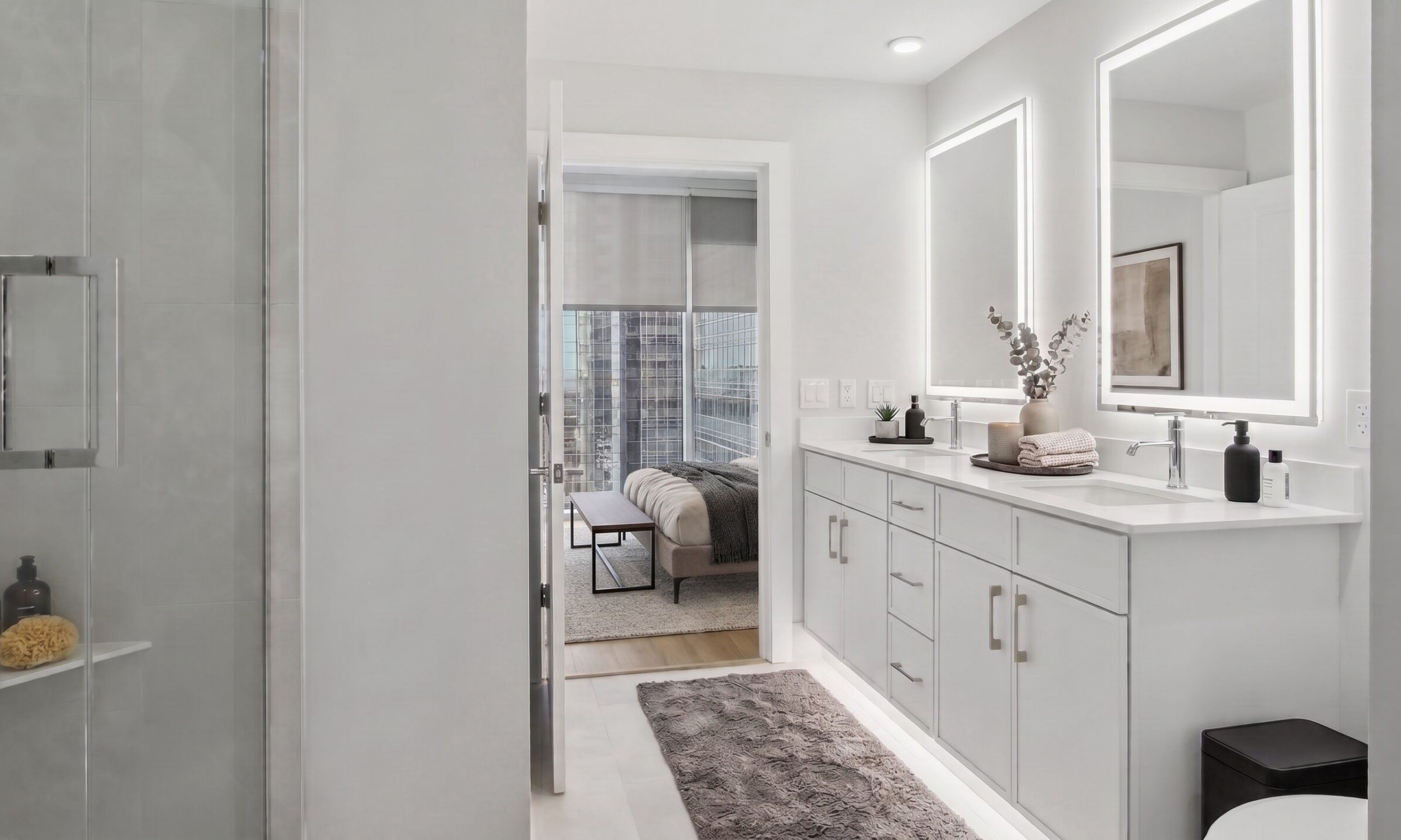 Modern bathroom with a double vanity and a glass shower, looking into a bedroom with city views.