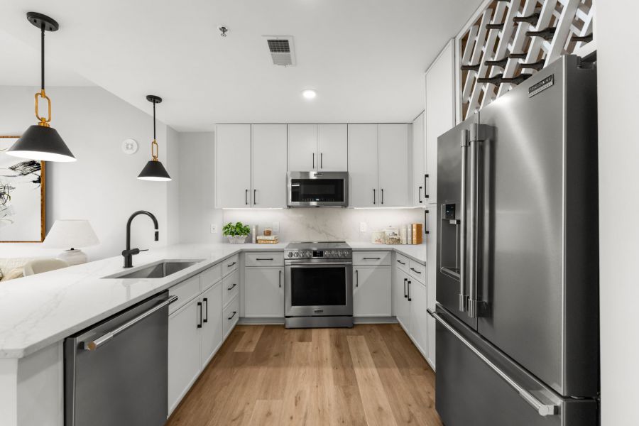 Modern kitchen with white cabinets, stainless steel appliances, black fixtures, and wood flooring. Marble countertops and pendant lights.
