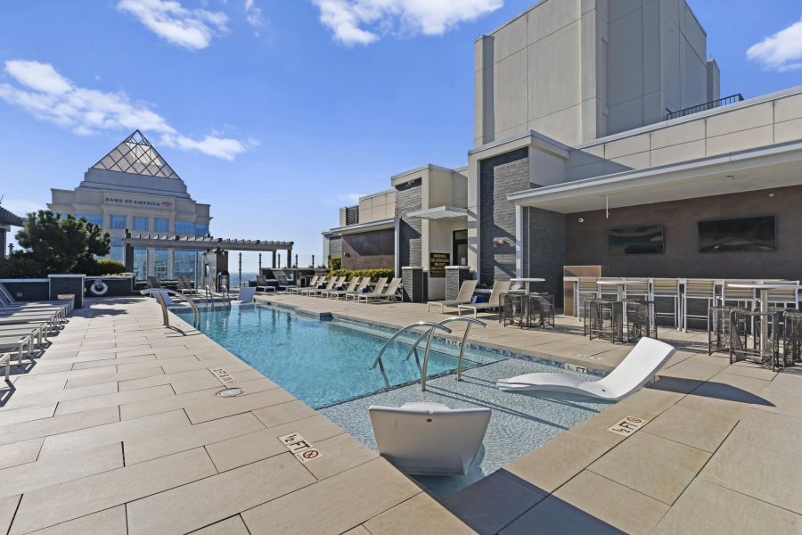 Rooftop pool with lounge chairs, shaded seating, and city buildings under a bright blue sky.