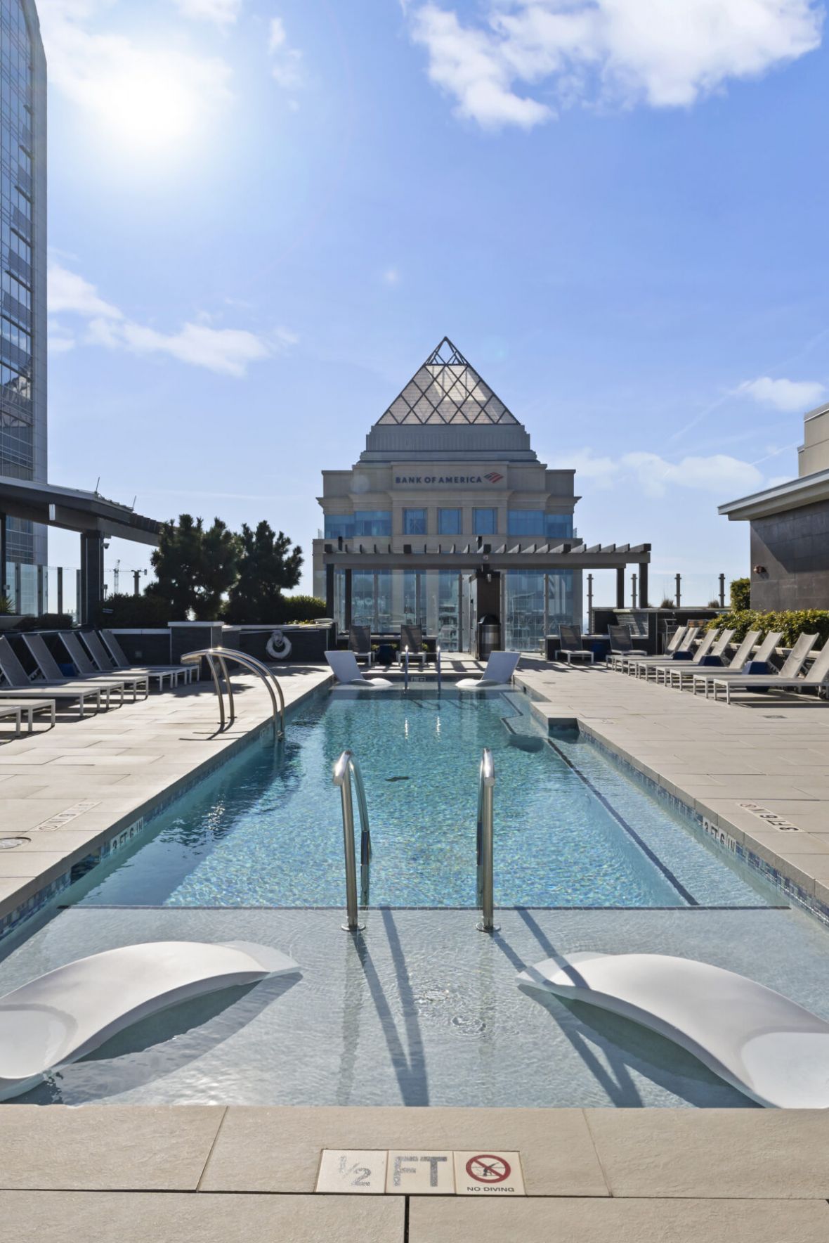 Rooftop pool with two submerged loungers, surrounded by chairs and tall buildings under a bright blue sky.