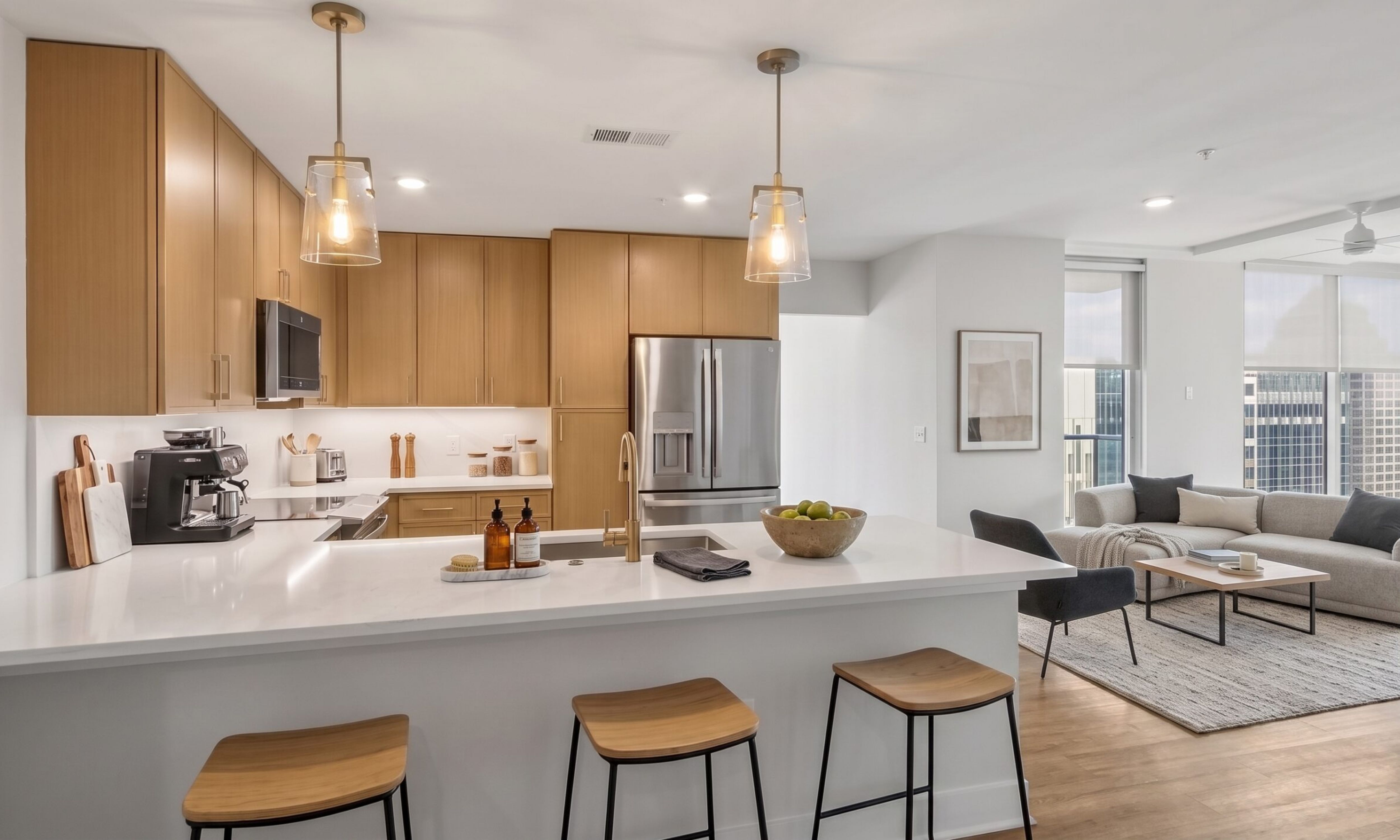 Modern kitchen with light wood cabinets, white countertops, and bar stools, opening into a living room with a sofa and floor-to-ceiling windows.
