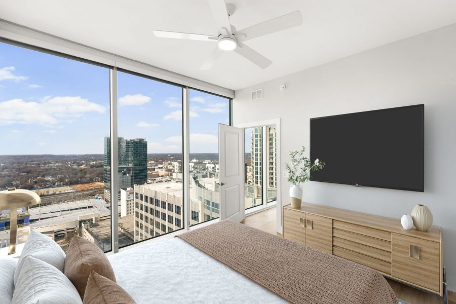 Modern bedroom with large windows, city views, a ceiling fan, bed, dresser, and a wall-mounted TV.