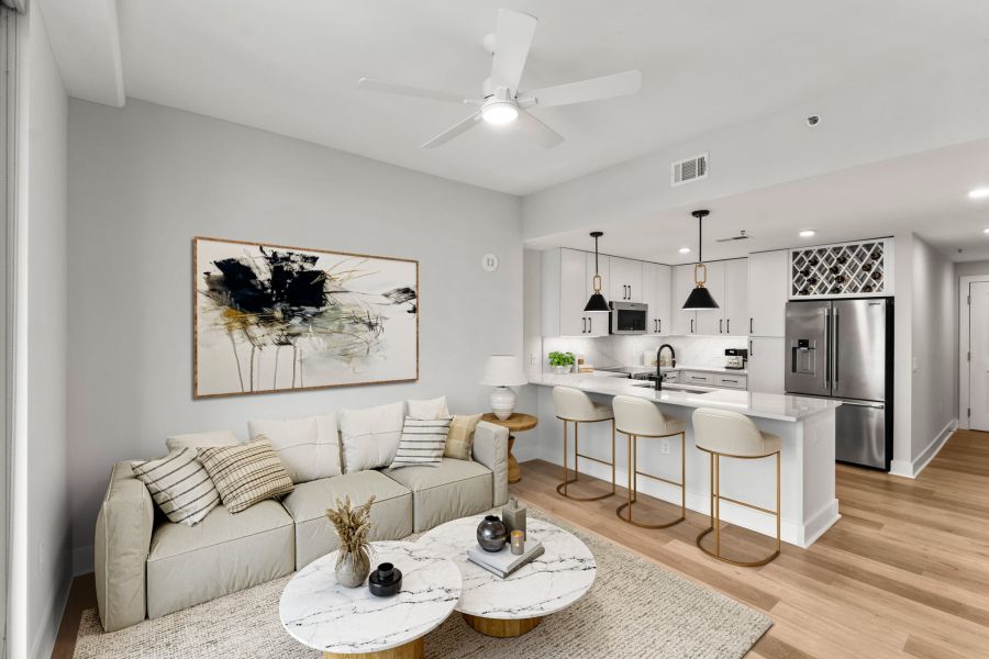 Modern open-concept living room and kitchen with beige sofa, marble tables, and white bar stools at a kitchen island.