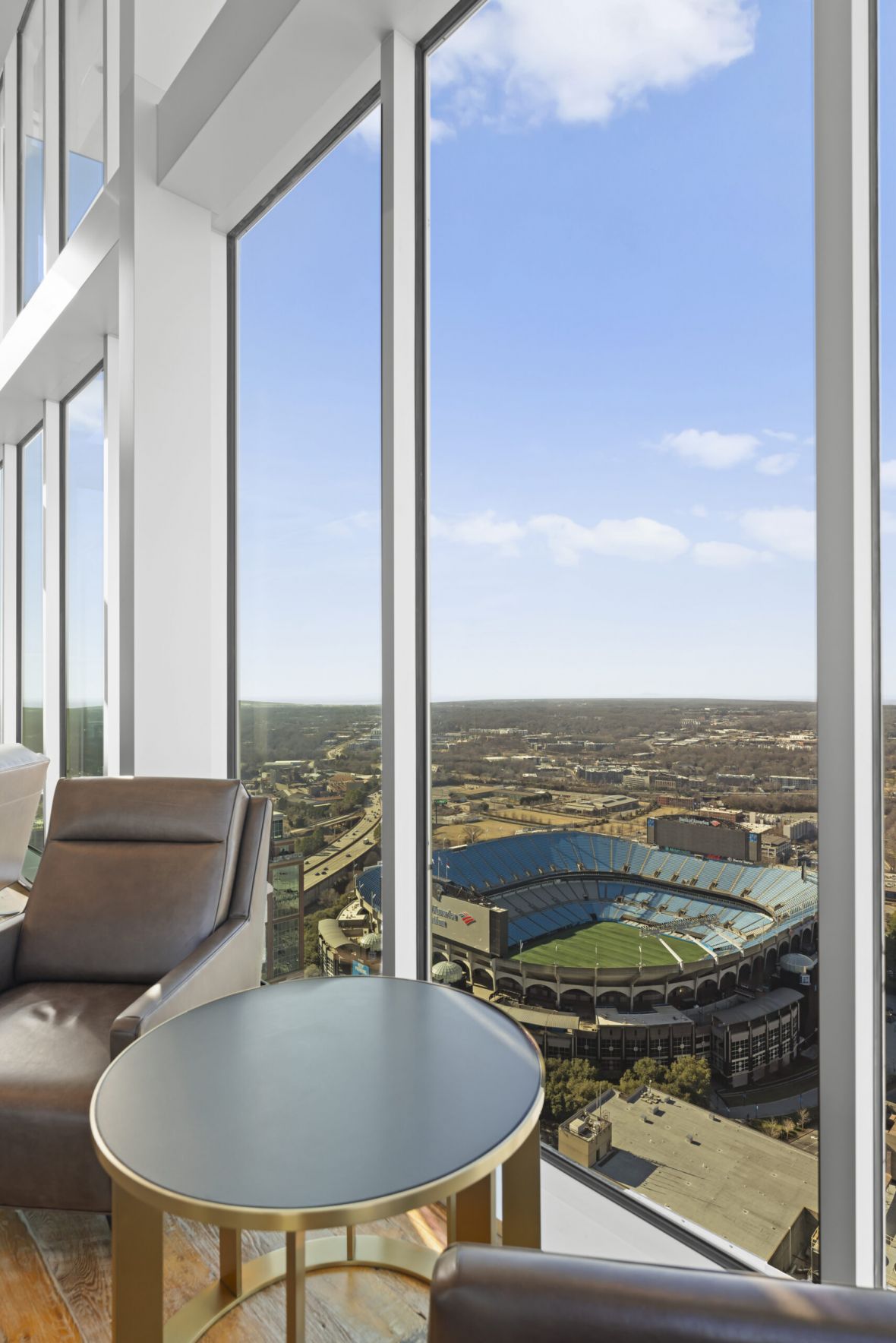 Modern lounge with leather chairs and round tables, overlooking a large football stadium through floor-to-ceiling windows.