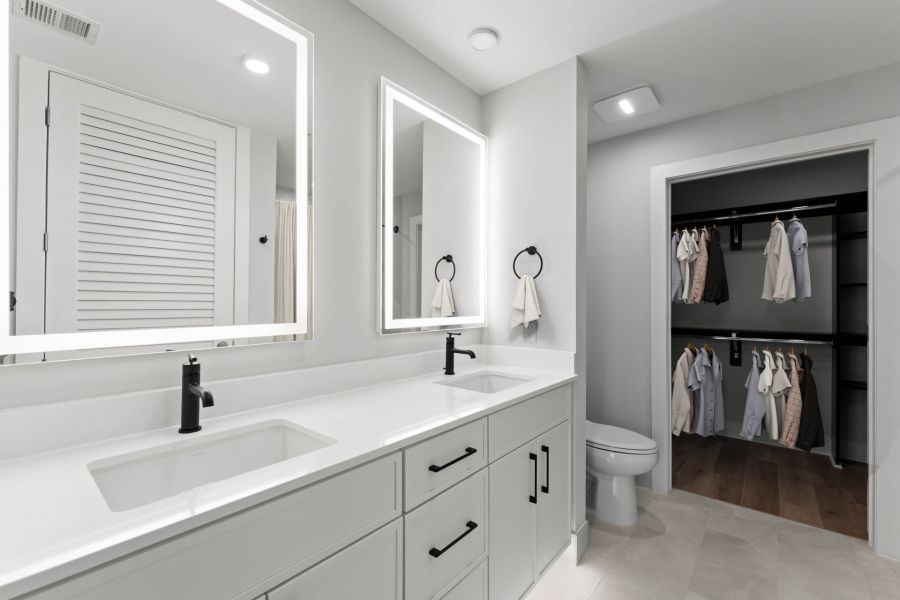 Modern bathroom with double sinks, large mirrors, white cabinets, and a walk-in closet with clothes hanging inside.