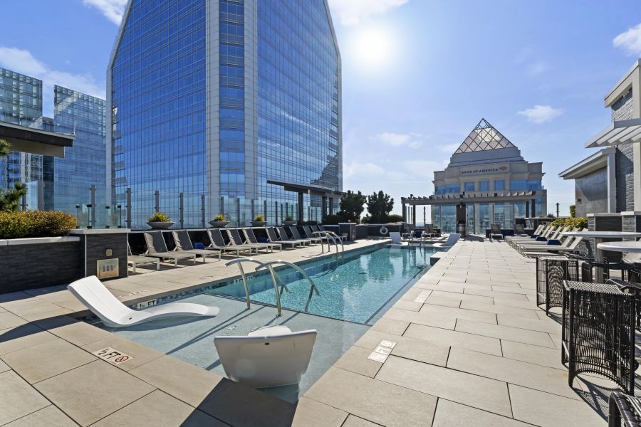 Rooftop pool surrounded by lounge chairs, with tall glass buildings and blue sky in the background.