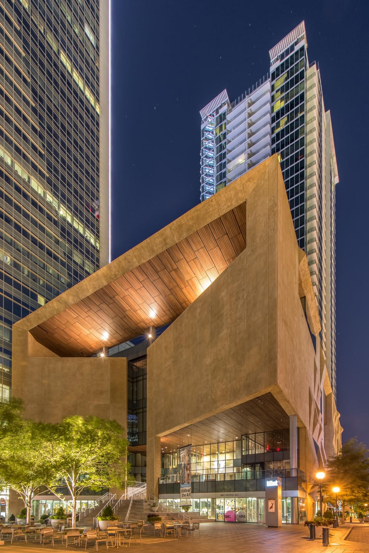 Modern city buildings and a unique angular structure lit up at night, with trees and outdoor seating in front.