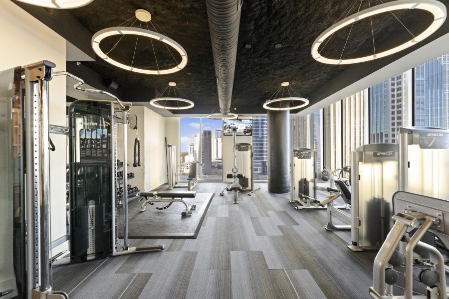Modern gym with exercise machines, large windows, city skyline view, and circular ceiling lights.